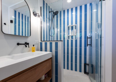 Modern primary bathroom featuring bold blue and white vertical striped tile, wall-mounted faucet, and oak vanity.
